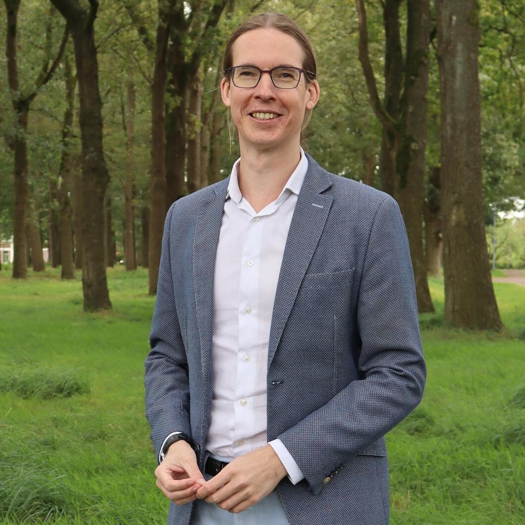 Wethouder Hans Kuipers draagt een grijze blazer met een witte blouse. Op de achtergrond staan wat bomen en gras