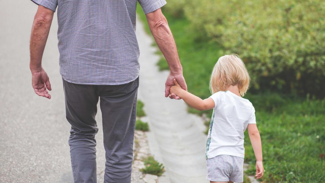 Een kind mee aan de hand van de vader. Op de achtergrond is groen gras te zien