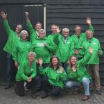 Op deze vrolijke groepsfoto zie je 11 vrouwen en mannen, allemaal gekleed in groene, GroenLinks jasjes. Ze zwaaien enthousiast naar de camera.. 