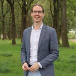Wethouder Hans Kuipers draagt een grijze blazer met een witte blouse. Op de achtergrond staan wat bomen en gras
