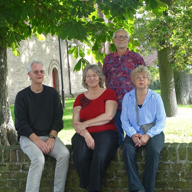 Groepsfoto van ons bestuur. Ze zitten op een stenen bankje, met op de achtergrond de kerk van Vries