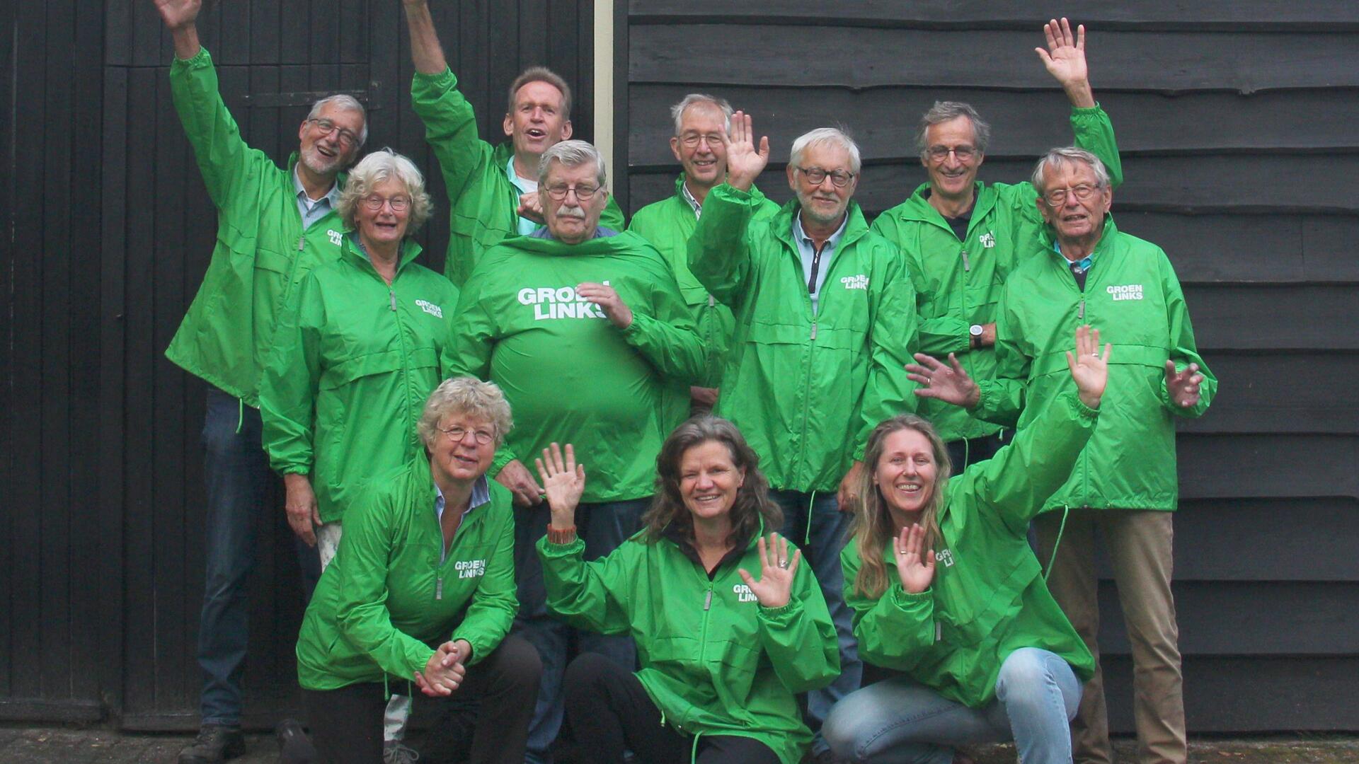 Op deze vrolijke groepsfoto zie je 11 vrouwen en mannen, allemaal gekleed in groene, GroenLinks jasjes. Ze zwaaien enthousiast naar de camera.. 