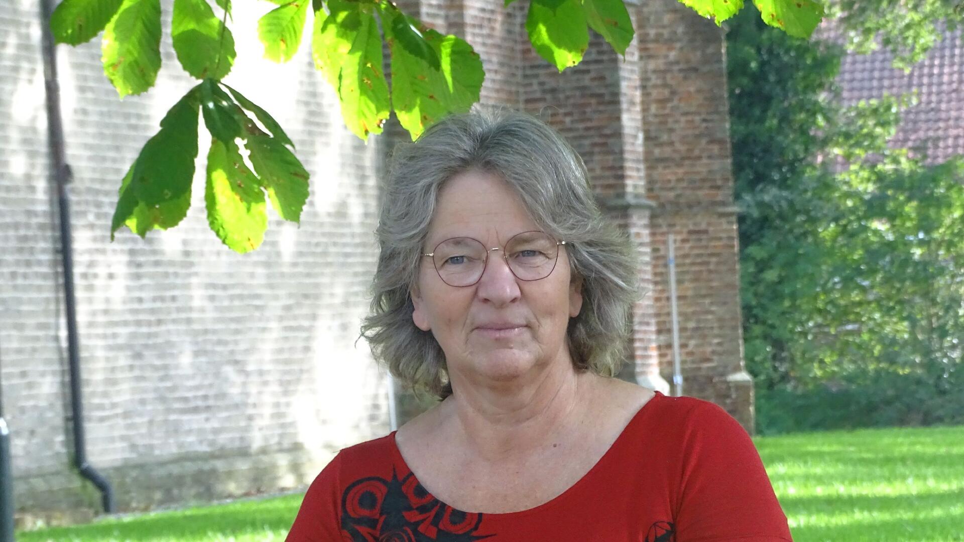 Lidwien Martens. Draagt een rood tshirt en zit voor een stenen kerk