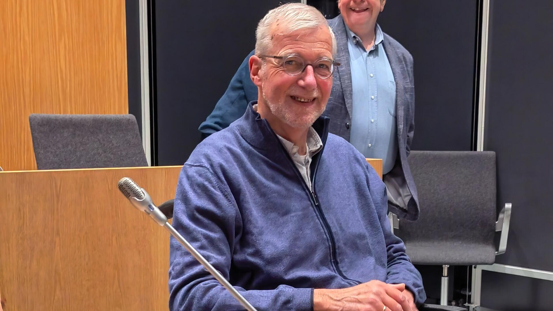 Raadslid Willem Nieman aanwezig bij de gemeenteraad. Hij heeft een blauwe blouse aan en zit in een handbewogen rolstoel