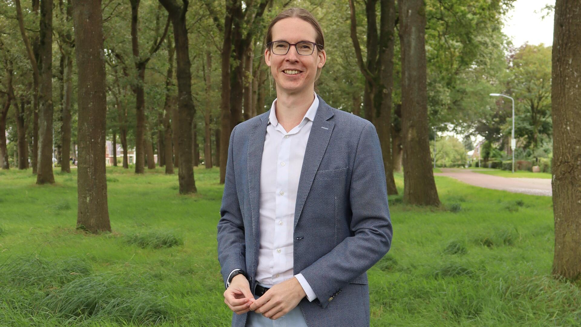 Wethouder Hans Kuipers draagt een grijze blazer met een witte blouse. Op de achtergrond staan wat bomen en gras