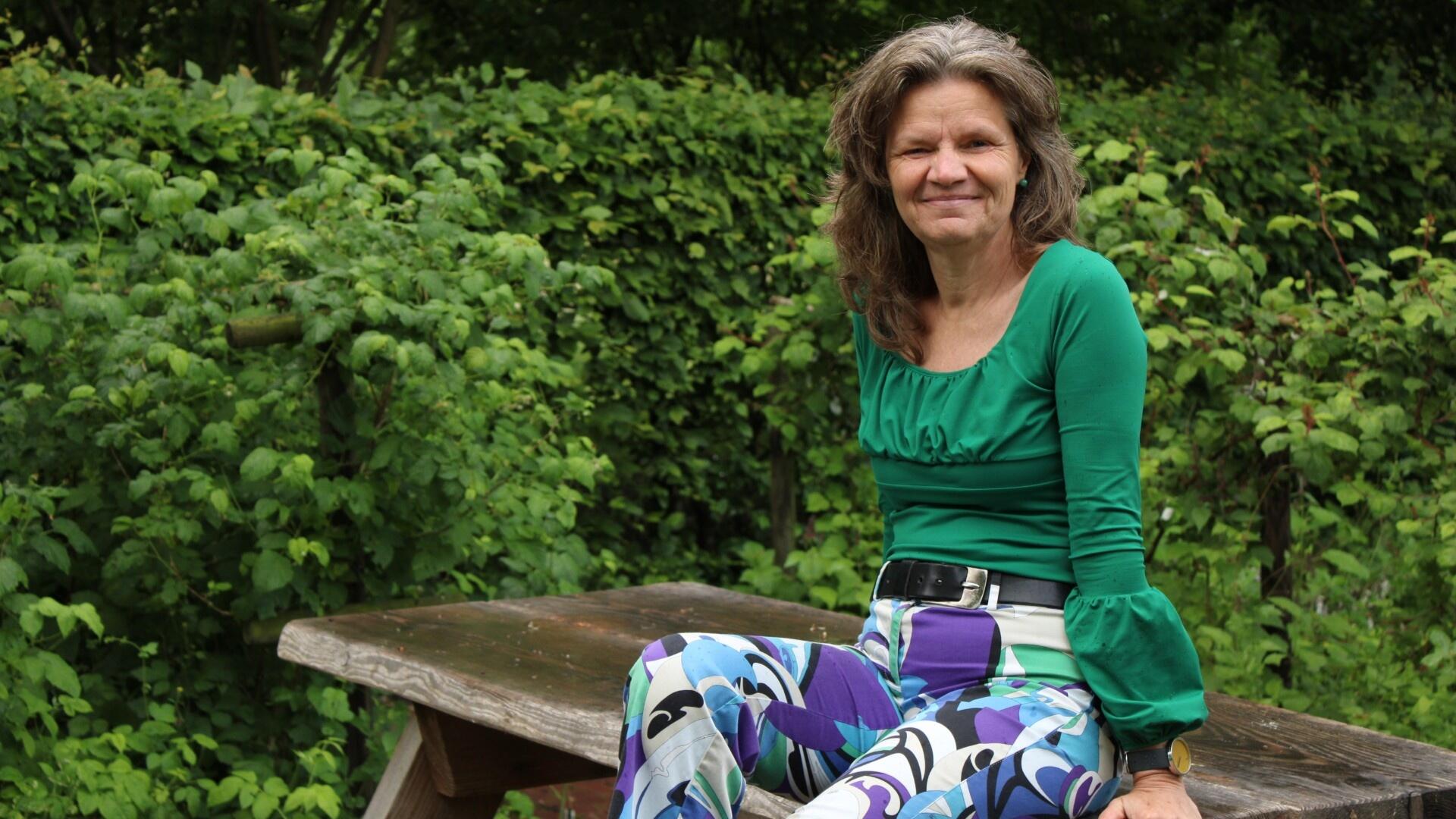 Gea Bijkerk, onze fractievoorzitter. Gea draagt een groen jasje en zit op een bankje voor wat groene struiken